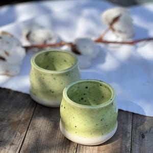 Può includere: Due piccole tazze in ceramica verde con smalto bianco macchiato. Le tazze sono su una superficie di legno.