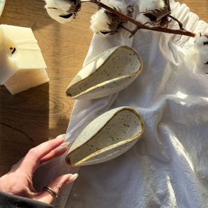 Op de afbeelding: Twee bespikkelde keramische lepelsteunen met een lichtbeige glazuur en een bruine rand. De lepelsteunen liggen op een witte doek, met katoenstelen en een brandende kaars op de achtergrond. De lepelsteunen zijn ongeveer 15 cm lang.