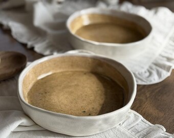May include: Two white ceramic bowls with a brown glaze on the inside. The bowls are sitting on a white ruffled tablecloth.