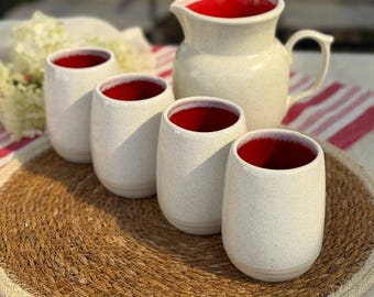 May include: A cream-colored ceramic pitcher and four matching tumblers with speckled exteriors and bright red interiors. The set is arranged on a woven placemat and a red and white striped cloth.
