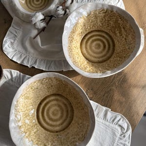 以下が含まれることがあります： 白い釉薬と茶色の斑点模様が施されたセラミック製のボウルが3つ。ボウルの中央には円形のデザインが施されています。