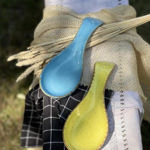 Puede incluir: Dos reposacucharas de cerámica, uno azul y otro amarillo, con esmalte moteado. Los reposacucharas están sobre una tela a cuadros negros y blancos.