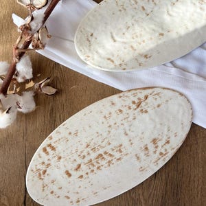 Puede incluir: Dos fuentes ovaladas de cerámica blanquecina con un diseño moteado marrón. Las fuentes están sobre un paño blanco sobre una superficie de madera. Una rama de algodón con vainas secas está en la esquina superior izquierda.