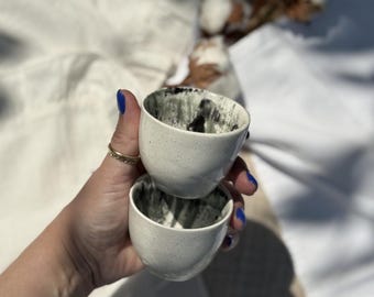 May include: Two small, speckled white ceramic cups with a dark gray interior. The cups are stacked on top of each other, held by a hand with blue nail polish. The cups have a unique, organic shape.