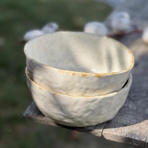 May include: Two white ceramic bowls with a speckled glaze and a brown rim. The bowls are stacked on top of each other and are sitting on a wooden surface.