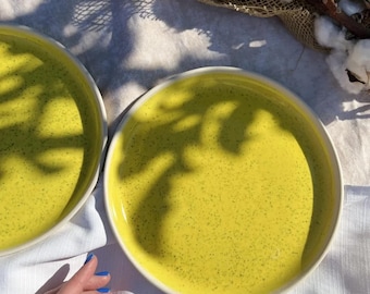 May include: Two yellow ceramic bowls with a speckled finish. The bowls are filled with a yellow liquid and are sitting on a white linen tablecloth. The bowls are illuminated by sunlight, casting shadows on the tablecloth.