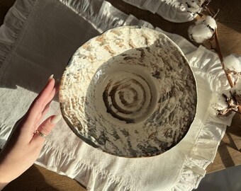 Può includere: Una grande ciotola in ceramica bianca con motivi a spirale marroni e neri. La ciotola è appoggiata su una tovaglia di pizzo bianca.