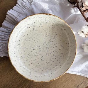 May include: A speckled white ceramic bowl with a brown rim. The bowl is set on a white cloth and a wooden surface. The bowl has a rustic, handmade appearance, suitable for serving food.