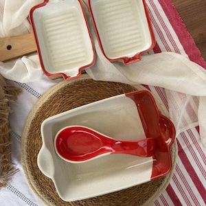May include: A collection of ceramic baking dishes and a spoon. Two rectangular baking dishes with red trim and a ribbed interior are visible. A third dish with a spoon is also present. The spoon and the trim on the dishes are red.