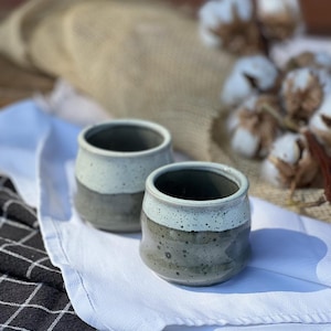 Peut inclure: Deux tasses en céramique avec un glaçage moucheté dans des tons de gris et de blanc. Les tasses sont posées sur un tissu blanc à motif à carreaux noirs et blancs.