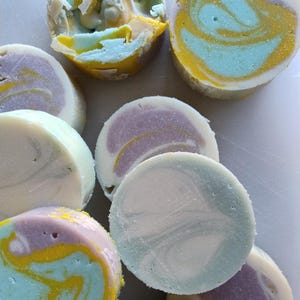 Op de afbeelding: Een close-up van verschillende ronde, swirl-vormige zepen in tinten blauw, geel en paars. De zepen zijn gerangschikt op een wit oppervlak.