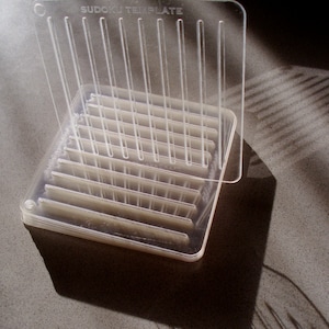 May include: A clear plastic Sudoku template with the words "Sudoku Template" printed on the top. The template is designed to help people solve Sudoku puzzles.