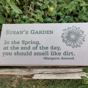May include: A light brown garden sign with the text "SUSAN'S GARDEN" and a quote by Margaret Atwood: "In the Spring, at the end of the day, you should smell like dirt."