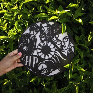 Puede incluir: Un reloj de pared circular en blanco y negro con recortes intrincados de tres personajes y las letras HP. El diseño incluye una esfera central con números y retratos estilizados de tres figuras, todo elaborado en un material plano y oscuro.
