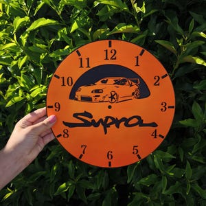 May include: A round orange wall clock featuring a black silhouette of a sports car above the word Supra in black script. The clock face displays black numerals from one to twelve and black tick marks for each hour, with a central hole for hands.