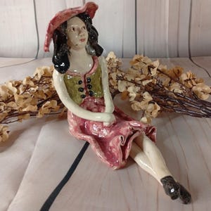 May include: A ceramic figurine of a seated figure in a pink dress and hat, with a green vest. The figure has black hair and black shoes. Dried flowers are in the background. The figurine is a decorative item.