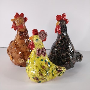 Puede incluir: Tres figuras de gallina de cerámica. Una es marrón, otra amarilla con un estampado floral y la tercera negra con motas rojas y marrones. Cada una tiene una cresta y una papada rojas. La gallina amarilla tiene una pajarita.