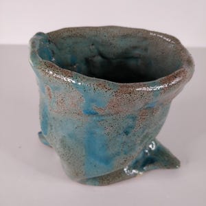 Könnte beinhalten: Eine handgefertigte Keramikvase mit einer einzigartigen, organischen Form. Die Vase hat eine gesprenkelte türkisfarbene Glasur mit braunen Sprenkeln und eine strukturierte Oberfläche. Der Rand ist uneben, was ihren handwerklichen Reiz unterstreicht. Für die Inneneinrichtung.