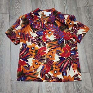 May include: A short-sleeved button-down shirt with a tropical print featuring orange, red, blue, and green leaves and flowers on a cream background. The shirt has a collar and a button closure.