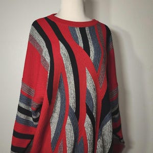 May include: Red sweater with a crew neck and long sleeves. The sweater features a bold design of vertical and diagonal stripes in black, gray, and blue. The ribbed knit cuffs and hem are red.