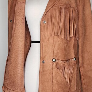 May include: A tan leather jacket with fringe detailing. The jacket features a collared neckline, button closures, and pockets with decorative studs. The fringe is along the sleeves, pockets, and chest.