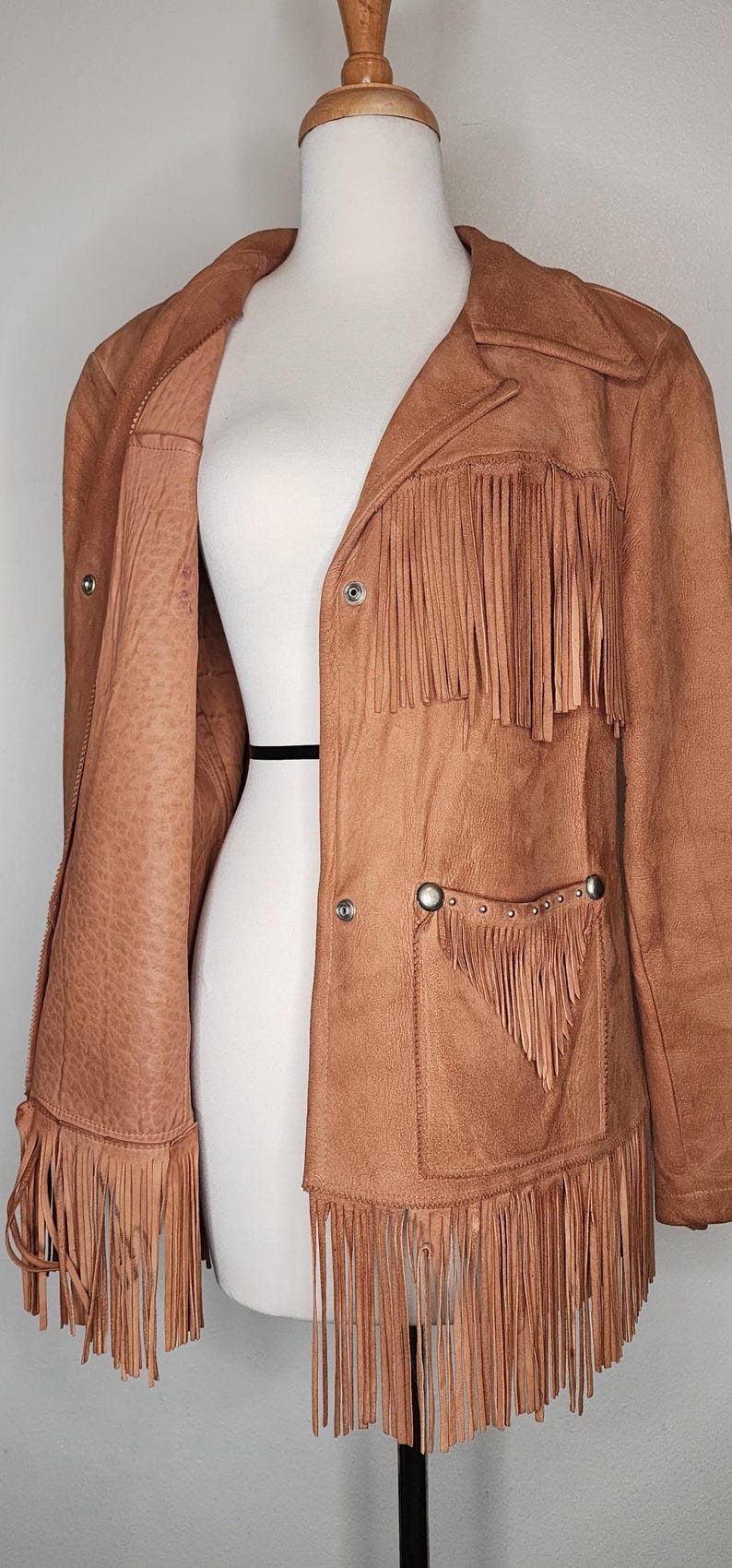 May include: A tan leather jacket with fringe detailing. The jacket features a collared neckline, button closures, and pockets with decorative studs. The fringe is along the sleeves, pockets, and chest.