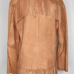 May include: Tan suede jacket with fringe detailing across the back and hemline. The jacket is displayed on a mannequin, showcasing its western-inspired design. The fringe adds a dynamic element to the garment.