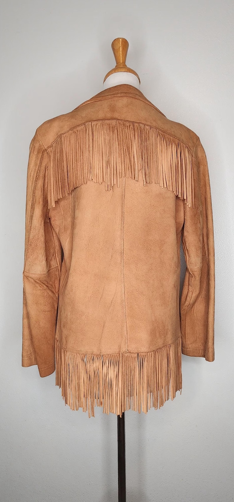 May include: Tan suede jacket with fringe detailing across the back and hemline. The jacket is displayed on a mannequin, showcasing its western-inspired design. The fringe adds a dynamic element to the garment.