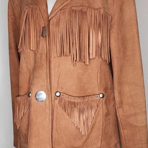 May include: A tan suede jacket with fringe detailing. The jacket features a notched collar, two front pockets, and large silver buttons. The fringe is along the front, pockets, and sleeves. A western-inspired garment.