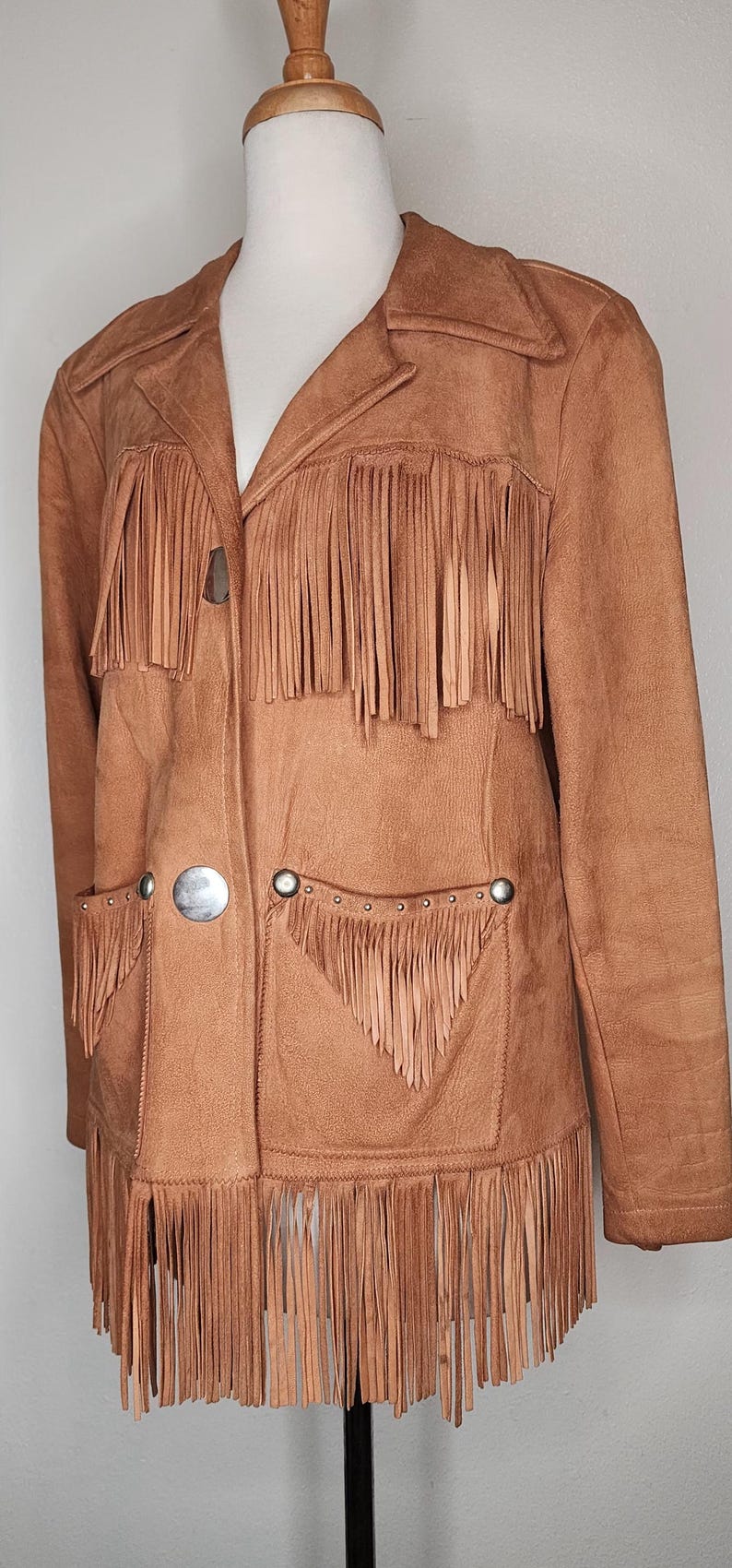 May include: A tan suede jacket with fringe detailing. The jacket features a notched collar, two front pockets, and large silver buttons. The fringe is along the front, pockets, and sleeves. A western-inspired garment.