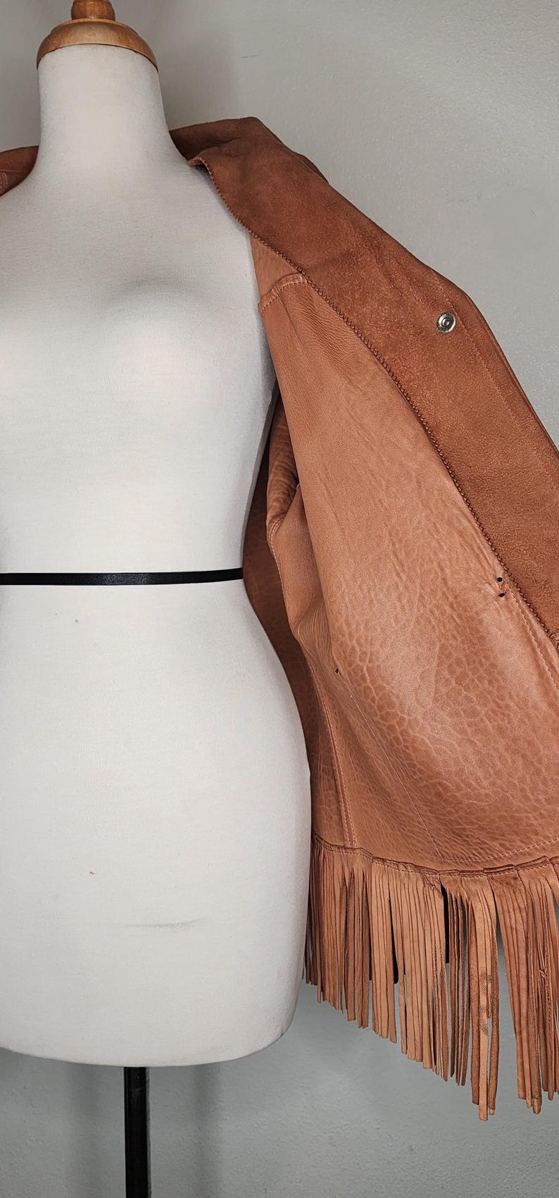 May include: A brown leather jacket with fringe detailing along the bottom hem. The jacket is displayed on a mannequin, showcasing its texture and design. The jacket has a button closure and a soft interior.