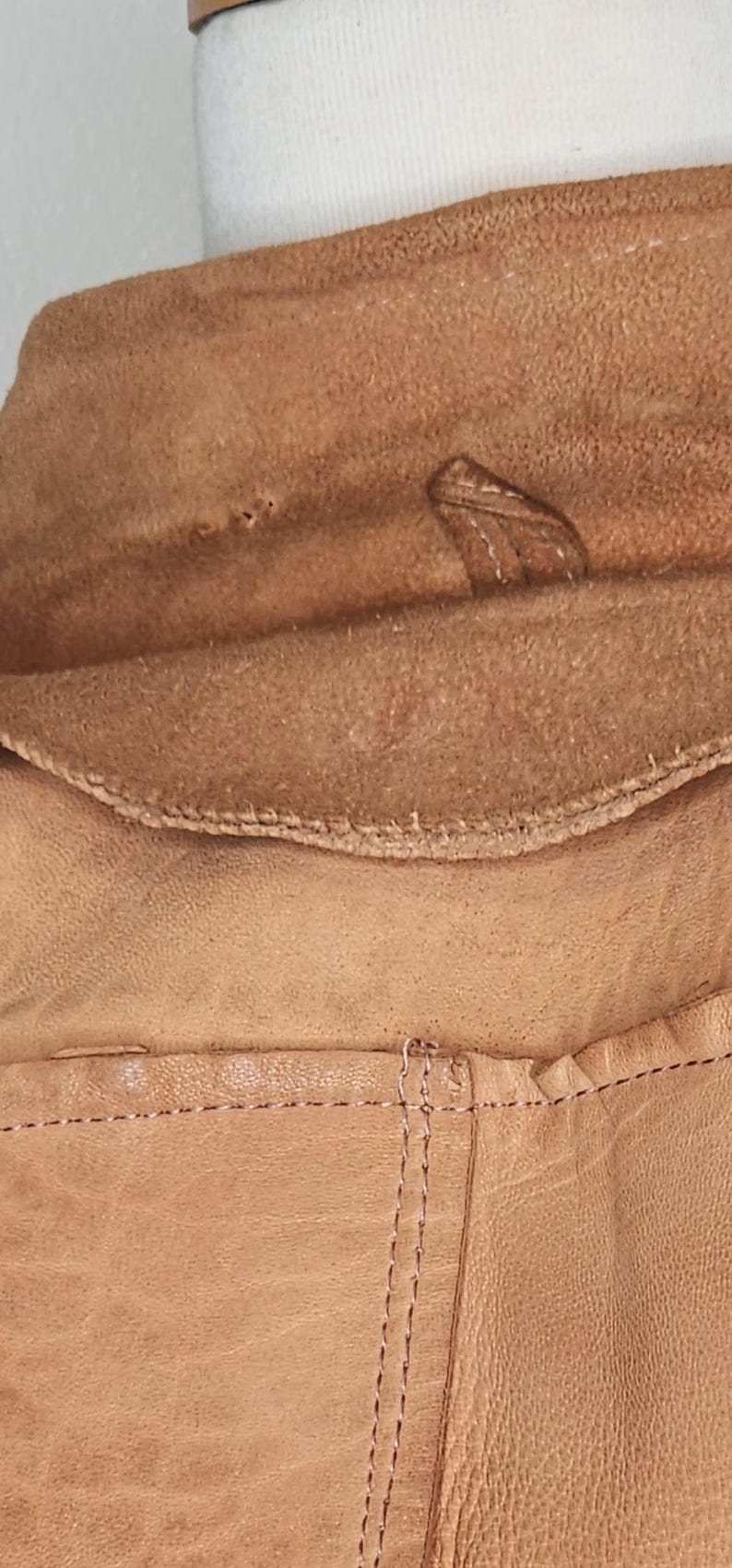 May include: Close-up of a tan leather jacket collar and lapel. The collar is folded over, revealing a soft, suede-like interior. Visible stitching and a small leather tab add detail to the design.