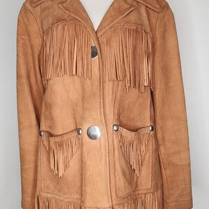 May include: A tan suede jacket with fringe detailing. The jacket features a collared neckline, two front pockets, and a button closure. The fringe is along the front, pockets, and sleeves. The jacket is displayed on a mannequin.