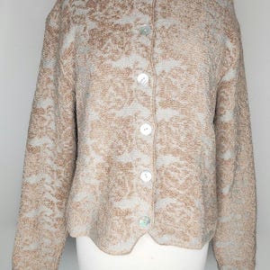 May include: A beige and light blue cardigan with a floral pattern. The long-sleeved sweater has a round neckline and is fastened with five white buttons. The garment is displayed on a white mannequin.