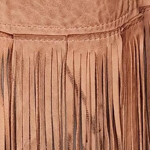 May include: Close-up of a tan leather garment with a fringe detail. The fringe is made of long, thin strips of leather, and the garment appears to be stitched together. The leather has a slightly distressed look.