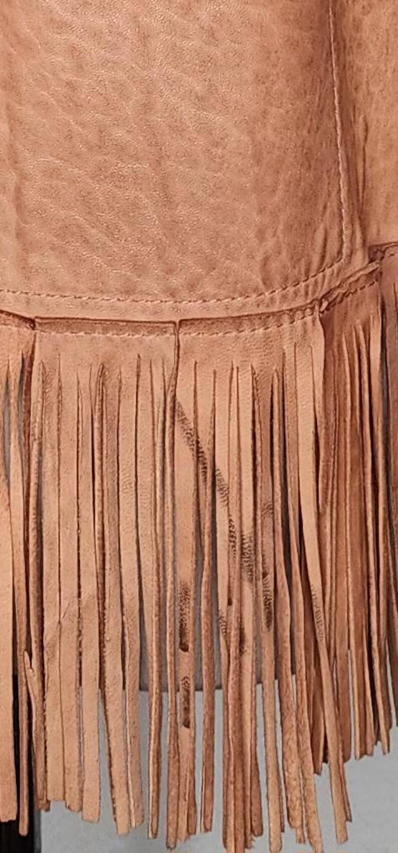 May include: Close-up of a tan leather garment with a fringe detail. The fringe is made of long, thin strips of leather, and the garment appears to be stitched together. The leather has a slightly distressed look.