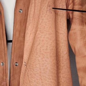 May include: A tan leather jacket with fringe along the bottom hem. The jacket has a button-up front and a textured appearance. The shoulders have a darker, more distressed look. The jacket is hanging on a hanger.
