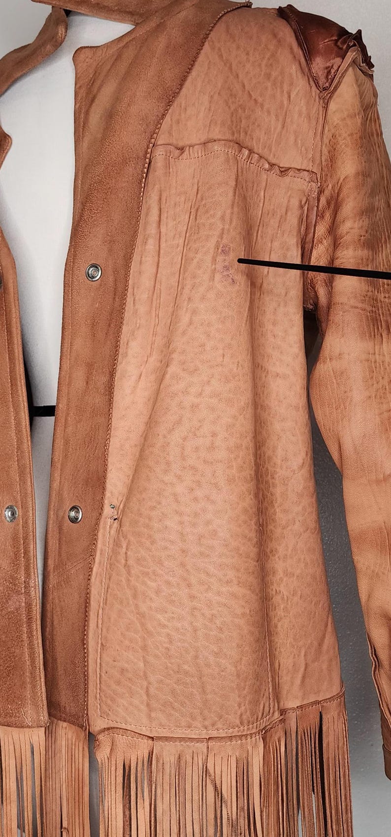 May include: A tan leather jacket with fringe along the bottom hem. The jacket has a button-up front and a textured appearance. The shoulders have a darker, more distressed look. The jacket is hanging on a hanger.