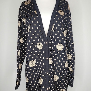 May include: A black cardigan sweater with a V-neck and long sleeves. The sweater features a pattern of small white dots and larger beige floral designs. It has gold-colored buttons down the front.