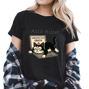 May include: Black t-shirt featuring a cartoon cat lying on a laptop with the words "PLEASE RESTART" above and "UNEXPECTED ERROR" on the screen. The cat is black and white. A fun, casual tee.