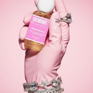 Peut inclure: Une bouteille d'huile parfum&eacute;e nourrissante Geminie Stardust est tenue dans une main gant&eacute;e de rose, orn&eacute;e de bagues et de bracelets en diamants. La bouteille a une &eacute;tiquette rose avec du texte blanc et un bouchon blanc, sur fond rose.