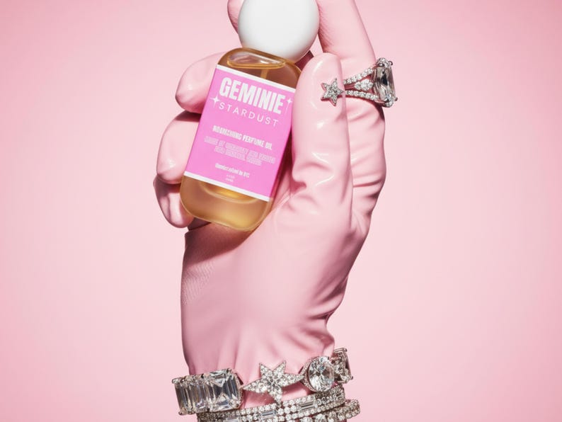 Peut inclure: Une bouteille d'huile parfum&eacute;e nourrissante Geminie Stardust est tenue dans une main gant&eacute;e de rose, orn&eacute;e de bagues et de bracelets en diamants. La bouteille a une &eacute;tiquette rose avec du texte blanc et un bouchon blanc, sur fond rose.