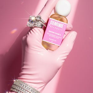 Peut inclure: Une main gant&eacute;e de rose tient une bouteille d'huile parfum&eacute;e Geminie Stardust. La main porte &eacute;galement une bague et un bracelet en diamants. La bouteille a une &eacute;tiquette rose avec du texte blanc. Le fond est rose uni.