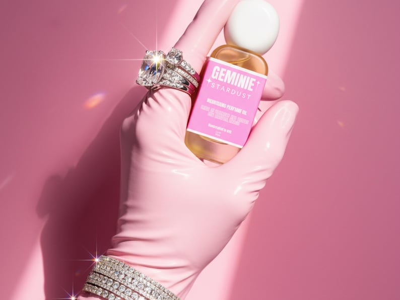 Peut inclure: Une main gant&eacute;e de rose tient une bouteille d'huile parfum&eacute;e Geminie Stardust. La main porte &eacute;galement une bague et un bracelet en diamants. La bouteille a une &eacute;tiquette rose avec du texte blanc. Le fond est rose uni.