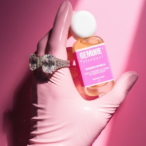 Peut inclure: Une bouteille d'huile parfum&eacute;e nourrissante Geminie Stardust avec une &eacute;tiquette rose est tenue par une main gant&eacute;e. La main porte &eacute;galement deux grandes bagues en diamant. Le fond est rose uni.
