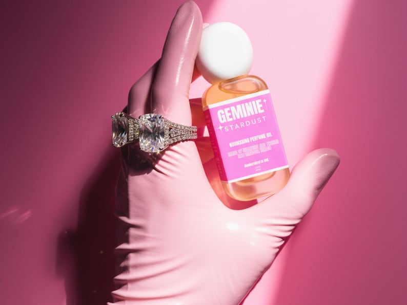 Peut inclure: Une bouteille d'huile parfum&eacute;e nourrissante Geminie Stardust avec une &eacute;tiquette rose est tenue par une main gant&eacute;e. La main porte &eacute;galement deux grandes bagues en diamant. Le fond est rose uni.