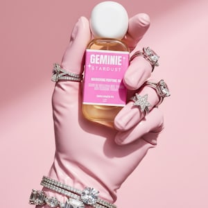 Peut inclure: Une bouteille d'huile parfum&eacute;e nourrissante Geminie Stardust est tenue par une main gant&eacute;e de rose orn&eacute;e de plusieurs bagues et bracelets en diamants. La bouteille a un bouchon blanc et une &eacute;tiquette rose avec du texte blanc.