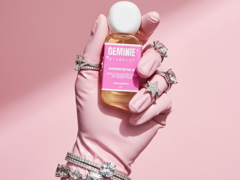 Peut inclure: Une bouteille d'huile parfum&eacute;e nourrissante Geminie Stardust est tenue par une main gant&eacute;e de rose orn&eacute;e de plusieurs bagues et bracelets en diamants. La bouteille a un bouchon blanc et une &eacute;tiquette rose avec du texte blanc.