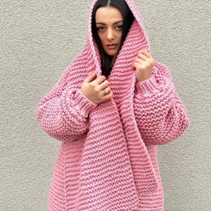 Könnte beinhalten: Ein rosa Strickcardigan mit Kapuze. Der Cardigan ist aus dickem Garn gefertigt und hat eine lockere Passform.