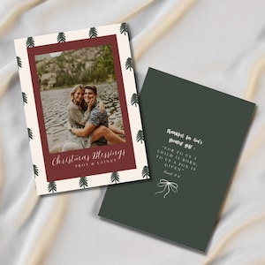 May include: Christmas greeting card with a photo of a couple embracing by a lake. The card has a burgundy border with a cream background and small green tree illustrations. The text "Christmas Blessings" is written on the front.
